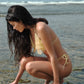 Woman in a yellow bikini crouching on a beach with ocean waves in the background